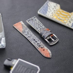 May include: Two watch straps made of patterned leather. One strap is gray, blue, and orange. The other strap is gray, blue, and white. Both straps have a silver buckle.