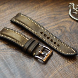 Può includere: Primo piano di un cinturino per orologio in pelle verde oliva invecchiata con cuciture color crema. Il cinturino presenta una fibbia argentata e diversi fori di regolazione. Il cinturino è su una superficie di legno.