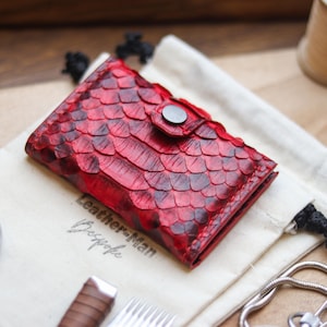 Könnte beinhalten: Ein rot-schwarzes Lederportemonnaie mit Schuppenmuster, gesichert mit einem silbernen Druckknopf. Das Portemonnaie liegt auf einem cremefarbenen Beutel mit dem Text "Leather-Man Bespoke". Weitere Artikel sind eine Stoppuhr und eine Garnrolle.