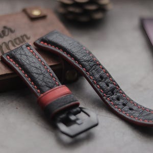 Pode incluir: Duas pulseiras de relógio de couro preto com costuras vermelhas. A pulseira à esquerda tem uma fivela e a pulseira à direita é plana. As pulseiras estão sobre uma superfície de couro marrom com o texto "Leather Man Handmade" visível.