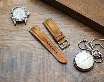 May include: A tan leather watch strap with visible stitching, a silver-toned metal buckle, and two keepers. The strap is positioned between a black-faced wristwatch with a silver case and a silver-toned pocket watch with a chain.
