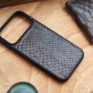 May include: A black phone case with a textured, snake-skin pattern. The case has a black border and a camera cutout. A small wallet with a similar pattern is in the background. The items are on a wooden surface.