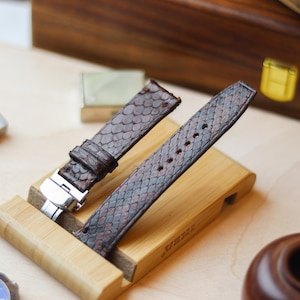 Könnte beinhalten: Ein Paar braune und graue Schlangenleder-Armbänder mit silberner Schließe, präsentiert auf einem Holzständer. Die Armbänder haben ein strukturiertes Muster und sind für eine Armbanduhr konzipiert. Der Hintergrund enthält Holzelemente.