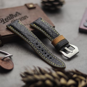 Pode incluir: Duas pulseiras de relógio de couro preto com costuras amarelas. Uma pulseira tem um inserto de couro castanho e uma fivela de metal. As pulseiras estão sobre uma superfície cinzenta.