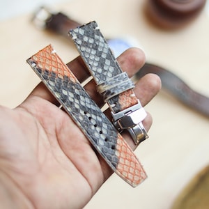 May include: Two-piece watch strap in patterned leather, featuring grey, black, and orange tones. The strap has a stainless steel buckle and is held in a hand. The watch strap is designed to be attached to a watch face.