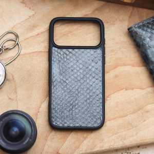 Könnte beinhalten: Eine schwarze Handyhülle mit Schlangenmuster, eine Stoppuhr, ein Objektiv und ein kleines Portemonnaie sind auf einer Holzoberfläche angeordnet. Die Handyhülle hat eine rechteckige Form mit einem Kameraausschnitt.