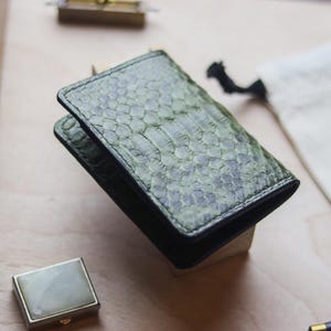 K&ouml;nnte beinhalten: Ein rechteckiger Kartenhalter mit einem strukturierten, olivgr&uuml;nen Schlangenledermuster mit sichtbaren Schuppen. Das Accessoire hat dunkle, gen&auml;hte Kanten und eine schlanke, gefaltete Konstruktion, die eine nat&uuml;rliche &Auml;sthetik aufweist.