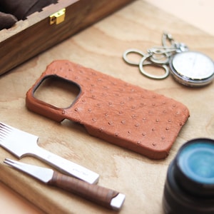 May include: A brown leather phone case with a textured surface, a silver pocket watch, and a silver and brown leather tool set are displayed on a wooden surface. A wooden box with a gold clasp is in the background.