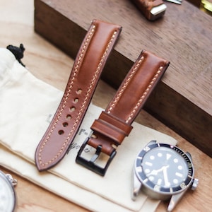 Pode incluir: Uma pulseira de relógio de couro marrom com costura creme e fivela preta é exibida ao lado de um relógio com mostrador preto e caixa prateada. Uma pequena bolsa creme e um cronômetro também estão na imagem.