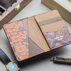 May include: A bi-fold wallet featuring a textured orange and black snakeskin pattern on the exterior panels, combined with smooth tan leather interior pockets. The interior includes a gold-stamped logo reading Leather-Mar Craft.