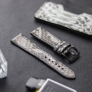Op de afbeelding: Twee grijze en witte slangenleren horlogebandjes met een zwarte gesp. De bandjes zijn gemaakt van echt leer en hebben een textuur.