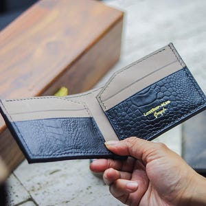 May include: A bi-fold wallet featuring a black exterior with a textured crocodile skin pattern and a smooth beige interior. The interior includes card slots and is embossed with the gold text Leather-Man Craft in a script and sans-serif font.