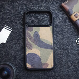 Può includere: Custodia per telefono nera con motivo mimetico nei toni del verde, marrone e beige. La custodia è circondata da un bordo nero. Altri oggetti includono un cronometro, un portafoglio in pelle e degli strumenti.