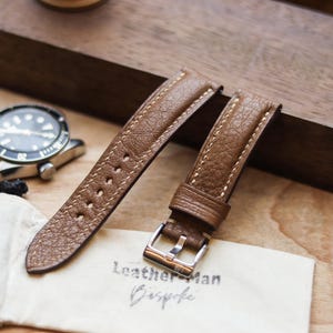 Könnte beinhalten: Zwei braune Leder-Uhrenarmbänder mit weißen Nähten und einer silbernen Schnalle. Die Armbänder werden auf einer Holzoberfläche mit einer Uhr und einem kleinen Beutel mit dem Text "Leather Man Bespoke" präsentiert.