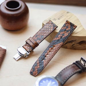 Könnte beinhalten: Nahaufnahme von Uhrenarmbändern mit Schlangenmuster in Braun, Schwarz und Orange. Ein Armband ist an einer Uhr mit blauem Zifferblatt befestigt. Weitere Lederwerkzeuge und eine Holzvase sind ebenfalls sichtbar.