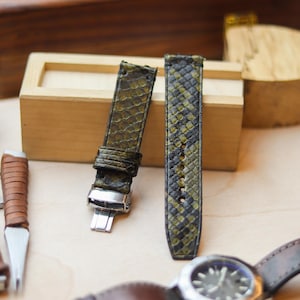 以下が含まれることがあります： オリーブグリーンと黒のヘビ柄の時計ストラップ2本、シルバーの留め金付き。 ストラップは明るい木製の表面に、茶色の革バンドの時計やその他の時計製造ツールと一緒に展示されています。 ストラップは新品です。