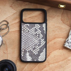 Può includere: Una custodia per telefono nera con un motivo a pelle di serpente nei toni del grigio e del bianco. La custodia è rettangolare con un ritaglio per la fotocamera e un bordo nero. Sono visibili anche un cronometro, un obiettivo e un piccolo oggetto in pelle.