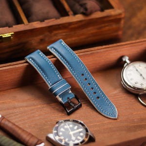 Pode incluir: Duas pulseiras de relógio de couro azul com costuras brancas e fivelas pretas são exibidas em uma superfície de madeira. Um cronômetro e um relógio também estão presentes, sugerindo um tema de cronometragem e acessórios.