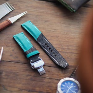 Könnte beinhalten: Zweifarbige, türkis-schwarze Leder-Uhrenarmbänder mit weißen Nähten und Uhrmacherwerkzeugen. Die Armbänder haben eine silberne Schnalle. Im Hintergrund ist ein Zifferblatt mit blauem Zifferblatt zu sehen.