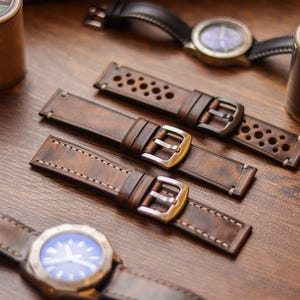 Könnte beinhalten: Nahaufnahme von braunen Lederarmbändern mit silbernen Schnallen, neben zwei Uhren und Garnrollen. Ein Armband hat ein perforiertes Design. Das Leder hat einen Used-Look, und die Uhren haben blaue Zifferblätter.