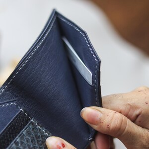 May include: A navy blue leather wallet featuring white contrast stitching along the edges. The interior displays a smooth navy blue lining with a dedicated slot containing a single silver-colored card. The exterior lower section has a textured finish.