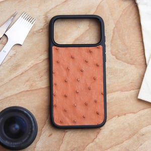 Può includere: Una custodia per telefono nera con un retro in pelle marrone rossiccio strutturato. La custodia è circondata da una cornice nera. Un obiettivo, strumenti in pelle e una piccola custodia sono su una superficie di legno.