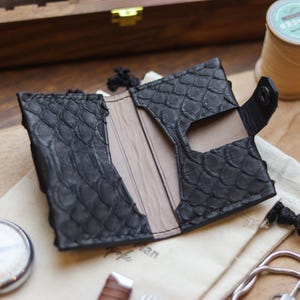 May include: Black leather wallet with a textured, scale-like pattern. The wallet is open, revealing a tan interior and card slots. A small black snap closure is visible. The wallet is on a light-coloured surface with other accessories.