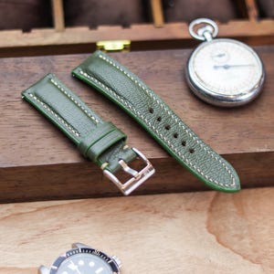 Pode incluir: Pulseira de relógio em couro verde com costura creme e fivela prateada. A pulseira é exibida em uma superfície de madeira, ao lado de um cronômetro e de um relógio.