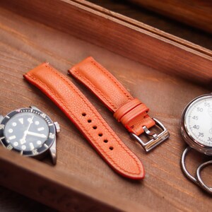 Pode incluir: Pulseiras de relógio de couro laranja com fivela prateada, um relógio com mostrador preto e um cronômetro são exibidos em uma bandeja de madeira. As pulseiras têm costuras visíveis.