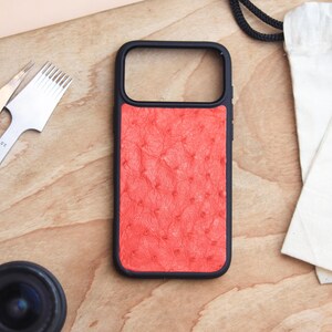 Può includere: Una custodia per telefono color corallo con una superficie strutturata, su uno sfondo di legno. La custodia ha una cornice nera. Nelle vicinanze ci sono strumenti per la lavorazione della pelle, un cronometro e una piccola lente.