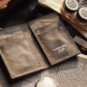 May include: Open, brown leather wallet with several card slots. The wallet has a distressed finish and the text "Leather-Man Design" is visible. The wallet is displayed open on a book and other accessories.