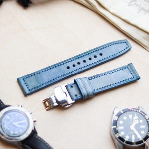 Può includere: Cinturini per orologi in pelle blu con cuciture bianche e chiusura argentata esposti su una superficie di legno. Sono visibili anche due orologi con cinturini neri e casse argentate. I cinturini sono di colore azzurro.