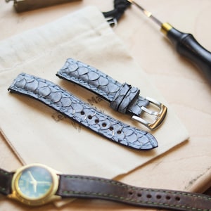 Könnte beinhalten: Zwei hellgraue Uhrenarmbänder mit Schuppenmuster, einer goldenen Schnalle und einem Leinensack werden auf einer Holzoberfläche präsentiert. Eine Uhr mit goldenem Gehäuse und braunem Lederarmband ist ebenfalls zu sehen, zusammen mit einem Lederwerkzeug.