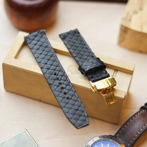 Könnte beinhalten: Zwei schwarze Uhrenarmbänder mit einem strukturierten, schuppenartigen Muster. Eines der Armbänder hat einen goldfarbenen Verschluss. Die Armbänder werden auf einer Holzkiste präsentiert, mit einer Uhr und anderem Zubehör im Hintergrund.