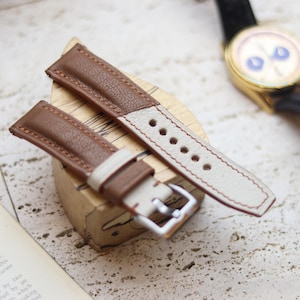 Può includere: Un cinturino per orologio in pelle marrone e crema con fibbia argentata. Il cinturino è bicolore, con pelle marrone sulla parte superiore e pelle crema sulla parte inferiore. Un orologio dorato con cinturino nero è sullo sfondo.