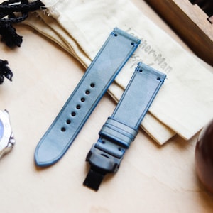 Könnte beinhalten: Zwei hellblaue Leder-Uhrenarmbänder mit schwarzen Verschlüssen und Nähten werden auf einer hellen Holzoberfläche präsentiert. Eine silberne Uhr mit blauem Zifferblatt und ein kleiner Kordelzugbeutel sind ebenfalls sichtbar. Die Armbänder sind neu.