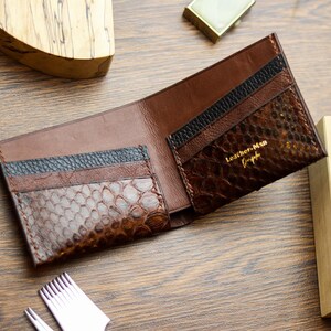 May include: A brown leather bifold wallet featuring textured snake-patterned panels, black leather card slots, and gold embossed text reading Leather-Man Gospoda. The interior includes multiple compartments for cards and currency storage.
