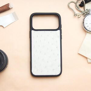 Può includere: Una custodia per telefono nera con un pannello bianco strutturato, forse in pelle di struzzo, è al centro dell'attenzione. Un cronometro, un obiettivo della fotocamera e strumenti per la lavorazione della pelle sono disposti attorno alla custodia su una superficie color pesca chiaro.