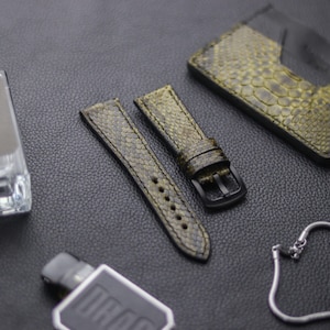 May include: Olive green watch straps with a textured, snakeskin-like pattern. The straps have a black buckle and are displayed with a matching cardholder on a dark surface. A silver chain and a small device are also visible.