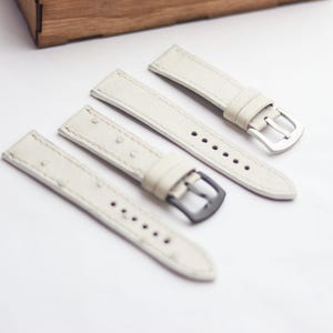 Könnte beinhalten: Vier cremefarbene Leder-Uhrenarmbänder mit silbernen und schwarzen Schnallen. Die Armbänder liegen auf einer weißen Oberfläche, mit einer Holzkiste im Hintergrund. Die Armbänder haben sichtbare Nähte und sind für eine Uhr bestimmt.