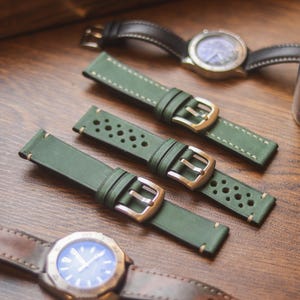 Può includere: Una vista dall'alto mostra diversi cinturini e orologi su una superficie di legno. Tre cinturini in pelle verde con fibbie argentate sono al centro. Sono visibili anche due orologi con quadranti blu e cinturini in pelle.