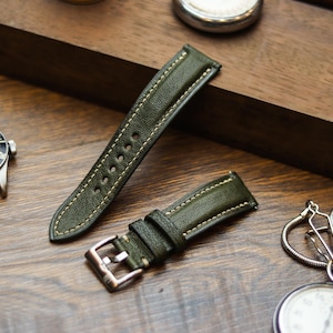 Pode incluir: Pulseiras de relógio em couro verde-oliva com costuras creme. As pulseiras são exibidas em uma superfície de madeira, ao lado de um relógio de pulso e um relógio de bolso. As pulseiras de relógio têm uma fivela prateada.