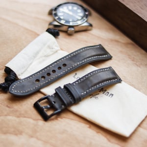 Può includere: Primo piano di un orologio e cinturini. L'orologio ha un quadrante nero e una cassa argentata. Due cinturini in pelle grigia con cuciture bianche sono esposti con un piccolo sacchetto. Il testo "The Man" è stampato sul sacchetto.