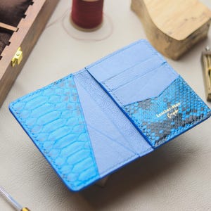 Può includere: Portafoglio in pelle blu chiaro aperto, con un motivo a pelle di serpente all'esterno. Il portafoglio ha più scomparti per carte e uno scomparto per banconote. Il marchio "Leather-Man" è visibile.