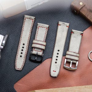 Könnte beinhalten: Vier cremefarbene Lederarmbänder mit roten Nähten und verschiedenen Schnallen. Die Armbänder sind im Used-Look mit Vintage-Optik. Der Hintergrund umfasst ein Lederetui, eine Kamera und eine Lederoberfläche.