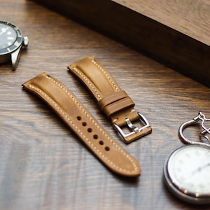 Pode incluir: Uma foto em close-up de um relógio, duas pulseiras de relógio de couro marrom e um cronômetro prateado em uma superfície de madeira. O relógio tem uma moldura preta e uma caixa prateada. As pulseiras são marrons claras com costuras creme.