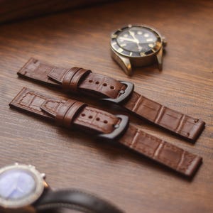 Può includere: Due cinturini per orologi in pelle marrone con fibbie nere sono esposti su una superficie di legno. È visibile un orologio da polso con quadrante nero e cassa argentata, insieme a un secondo orologio. È presente anche una bobina di filo.