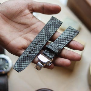 Puede incluir: Correas de reloj grises y blancas con estampado de piel de serpiente y cierre plateado. Las correas se sostienen en una mano, con un reloj visible en el fondo. Las correas miden aproximadamente 20 cm de largo.