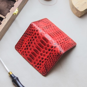 May include: A vibrant red bifold wallet featuring a distinct, raised snakeskin-like texture. The wallet is folded in a semi-open position, showcasing the detailed scale pattern across its exterior surface and the clean, stitched edges of the leather.