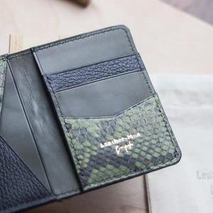K&ouml;nnte beinhalten: Ein aufklappbares Portemonnaie aus einer Kombination von gr&uuml;nem Leder mit Schlangenhautstruktur, glattem schwarzem Leder und dunkelblauem genarbtem Leder. Das Innere enth&auml;lt mehrere Kartenf&auml;cher und ist mit dem goldenen Text Leather-Man Bespoke gepr&auml;gt.